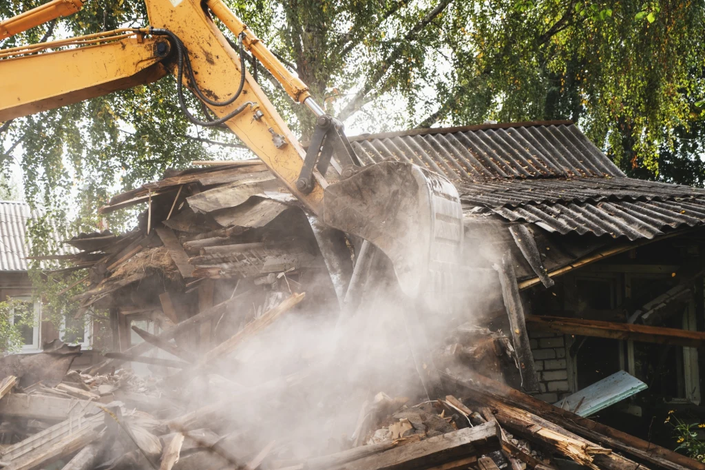 Koparka burząca stary drewniany budynek, w powietrzu unoszą się chmury pyłu, dach z blachy jest częściowo zawalony, wokół zniszczone elementy konstrukcji oraz gałęzie drzew w tle.