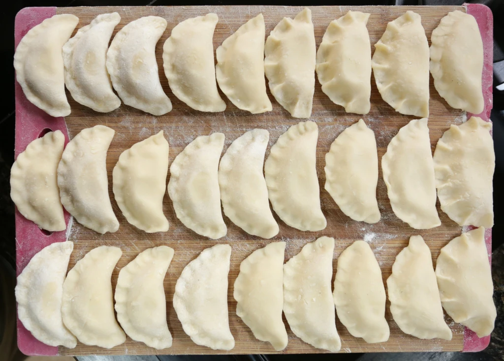 Pierogi to nieodłączny element wigilijnej kolacji Rządki surowych pierogów ułożonych równomiernie na drewnianej desce do krojenia, obsypanej lekko mąką, pierogi mają półokrągły kształt i są gotowe do gotowania.