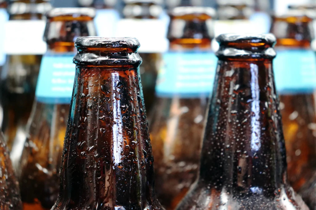 Zbliżenie kilku mokrych, szklanych butelek piwa ustawionych obok siebie, z wyraźnie widocznymi kroplami wody na ich szyjkach. Dominują brązowe kolory szkła i niebieskie etykiety w tle.