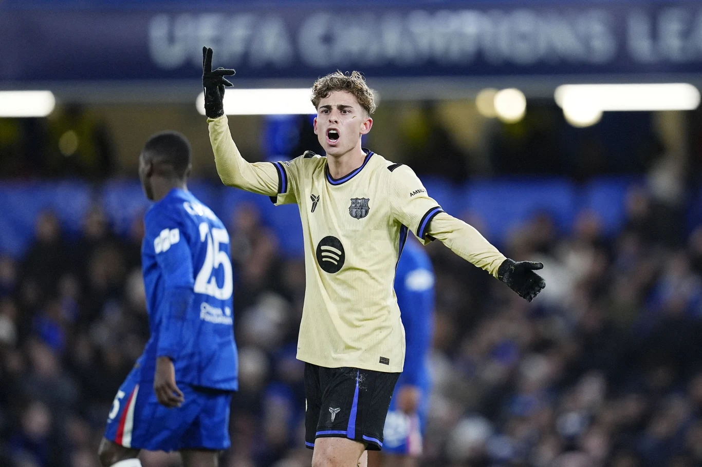 Piłkarz drużyny w jasnej koszulce gestykuluje obiema rękami podczas meczu na stadionie, z tyłu widoczny inny zawodnik w niebieskim stroju oraz rozmazany tłum kibiców.