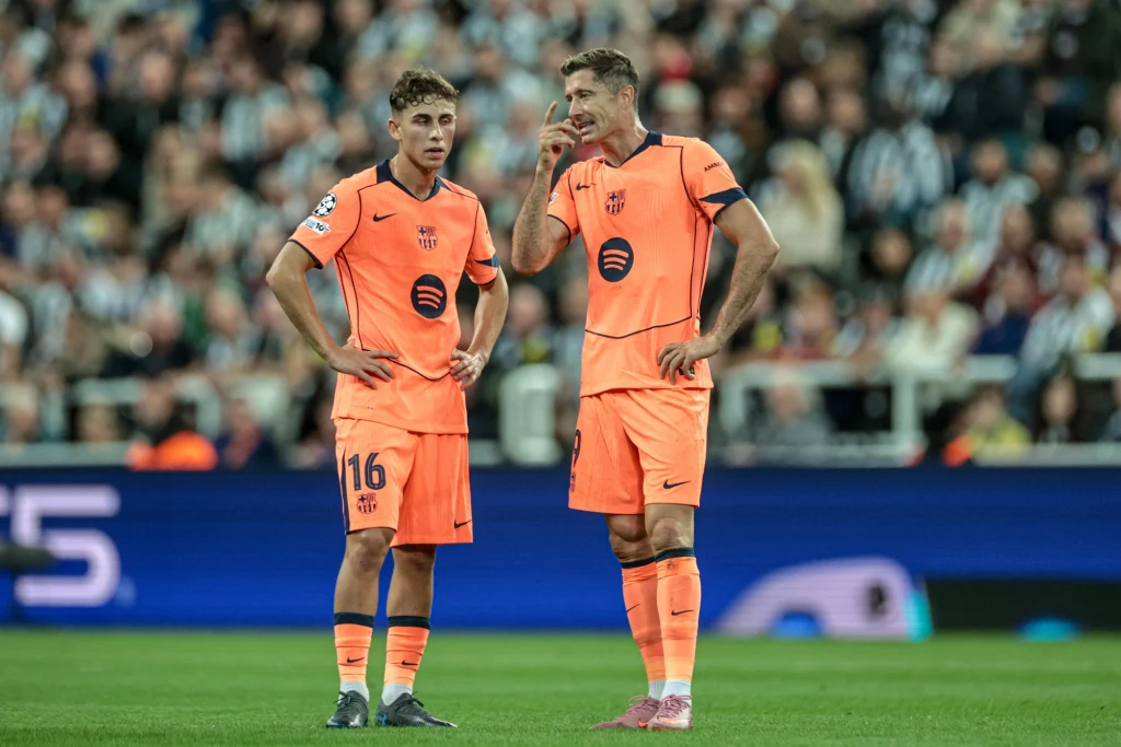 Dwóch piłkarzy w pomarańczowych strojach drużyny FC Barcelona stoi na murawie stadionu podczas meczu piłkarskiego. W tle widoczna publiczność oraz murawa boiska.