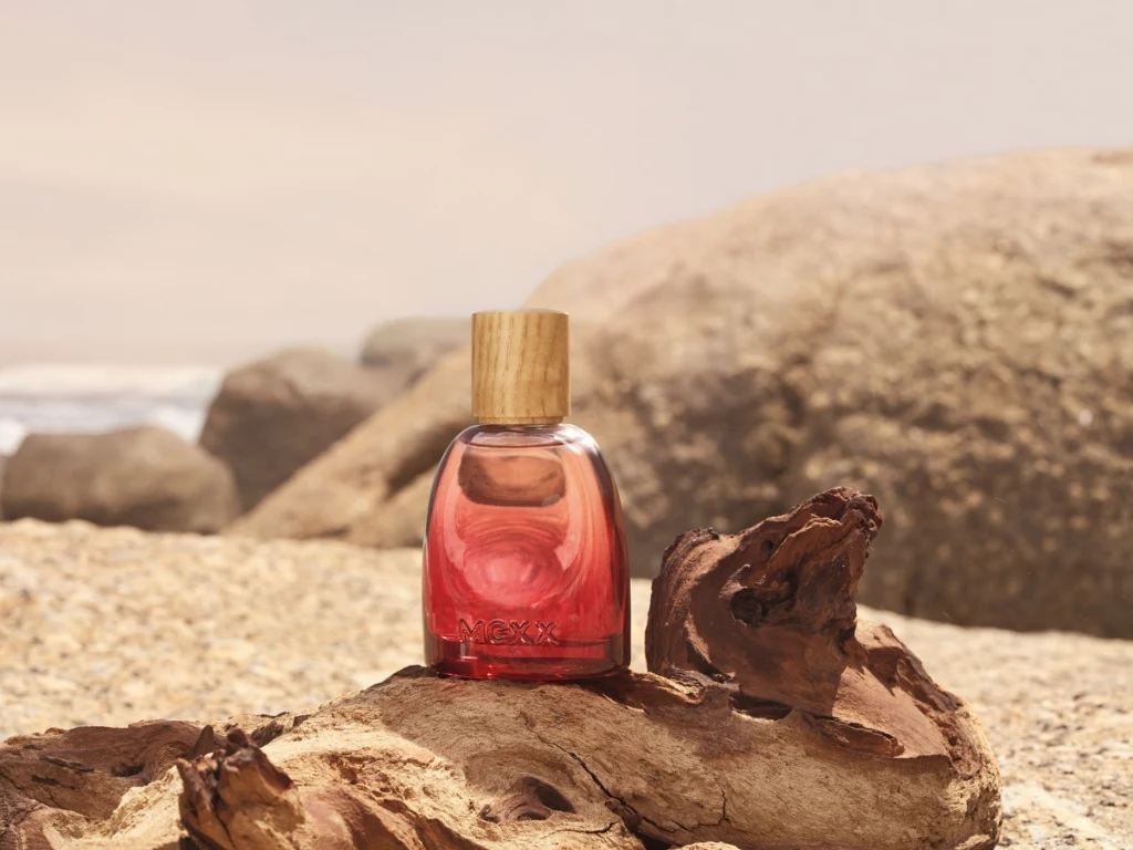 To perfumy, które kojarzą się z ciepłem, elegancją i dają uczucie przytulności Flakon czerwonych perfum z drewnianą zakrętką ustawiony na kawałku suchego drewna, na tle piasku oraz dużych skał w ciepłym, naturalnym świetle.