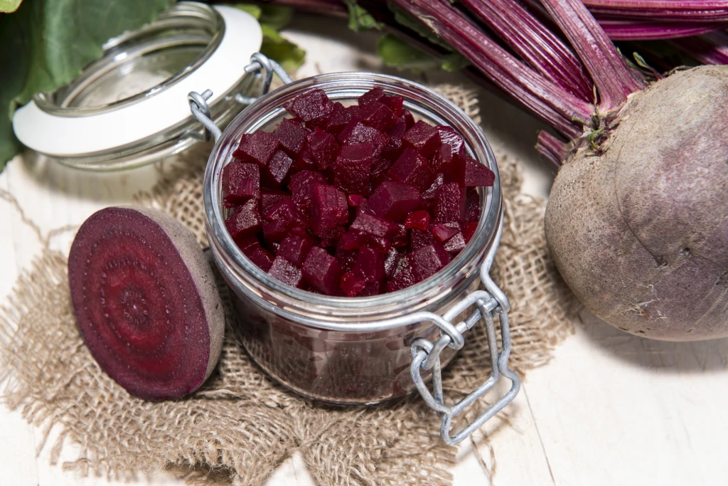 Kawałki czerwonych buraków w zalewie umieszczone w szklanym słoiku, obok przekrojony oraz cały surowy burak na jutowym materiale, w tle widoczne liście buraków.