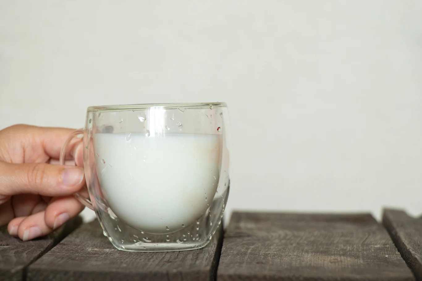 Szklanka przezroczysta wypełniona mlekiem, ustawiona na drewnianym stole, trzymana z boku przez dłoń.