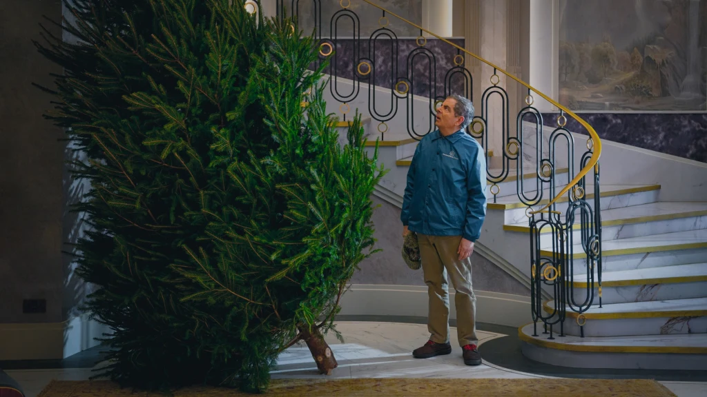 Mężczyzna w niebieskiej kurtce stojący przed dużą choinką w przestronnym wnętrzu domu, na tle eleganckich schodów z metalową balustradą.