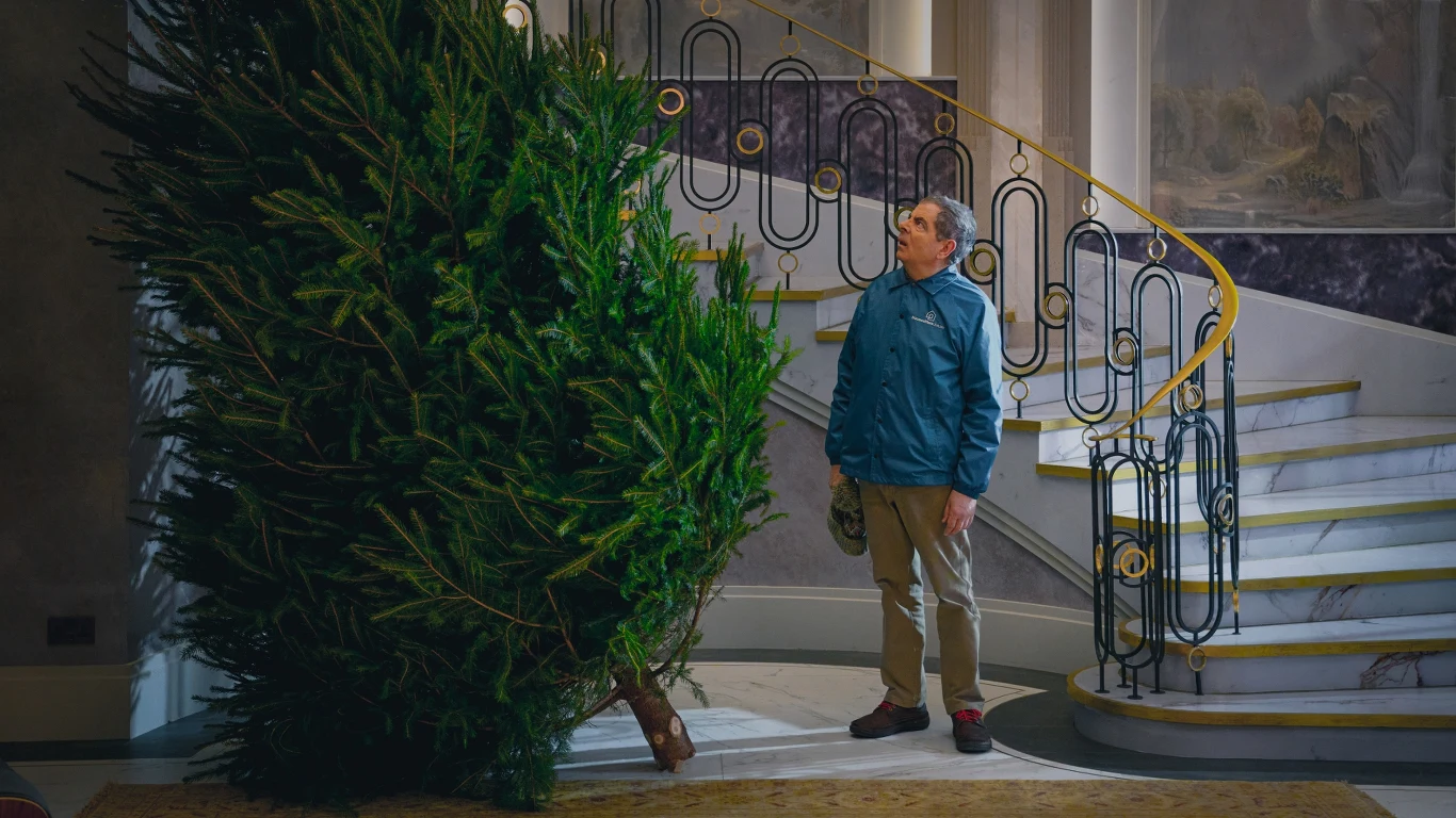 Mężczyzna w niebieskiej kurtce stojący przed dużą choinką w przestronnym wnętrzu domu, na tle eleganckich schodów z metalową balustradą.