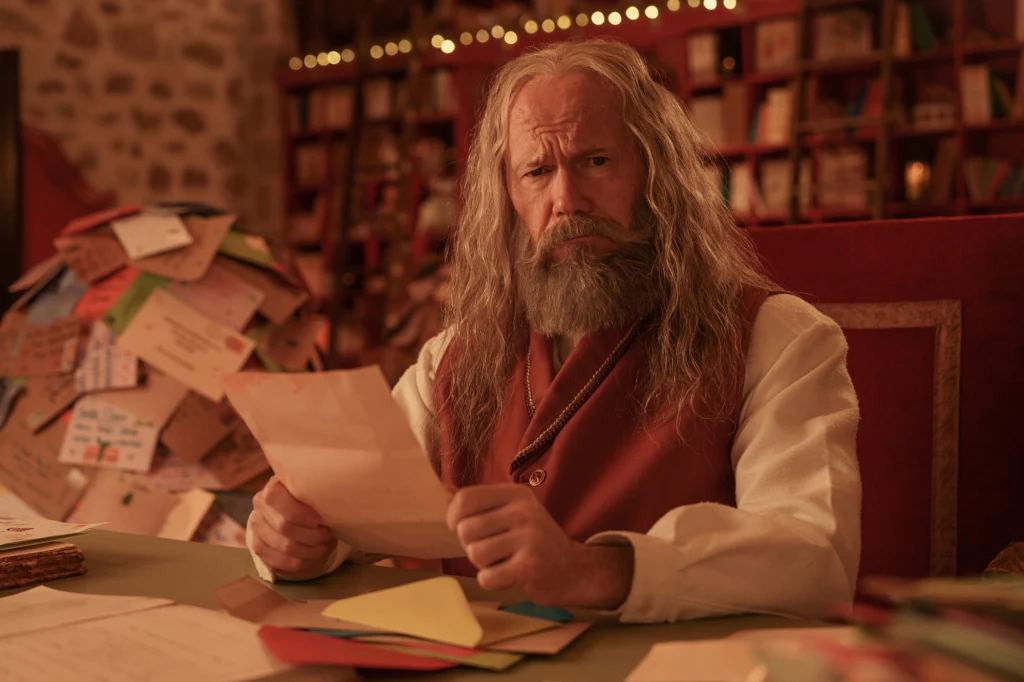 Starszy mężczyzna z długą siwą brodą i włosami, ubrany w kamizelkę i koszulę, siedzi przy stole pełnym kolorowych kopert i listów, trzymając jeden z nich w rękach, w tle półki pełne książek oraz stosy listów.