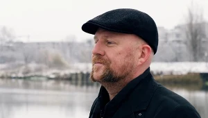 Mężczyzna w czapce z daszkiem patrzy w dal, tło stanowi zimowy krajobraz z rzeką i zaśnieżonymi drzewami.