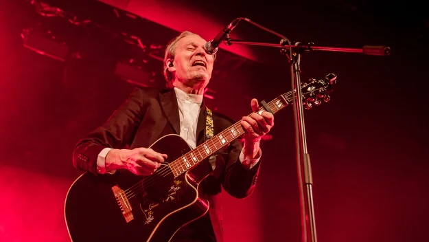 Mężczyzna w garniturze grający na gitarze akustycznej i śpiewający do mikrofonu podczas koncertu, scena oświetlona intensywnym, czerwonym światłem.