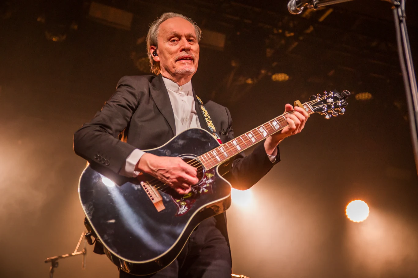 John Porter "Helicopters '45" w Warszawie Mężczyzna w średnim wieku w eleganckim garniturze gra na czarnej gitarze akustycznej podczas koncertu scenicznego, oświetlony przez jasne reflektory, z widocznym wyrazem skupienia na twarzy.