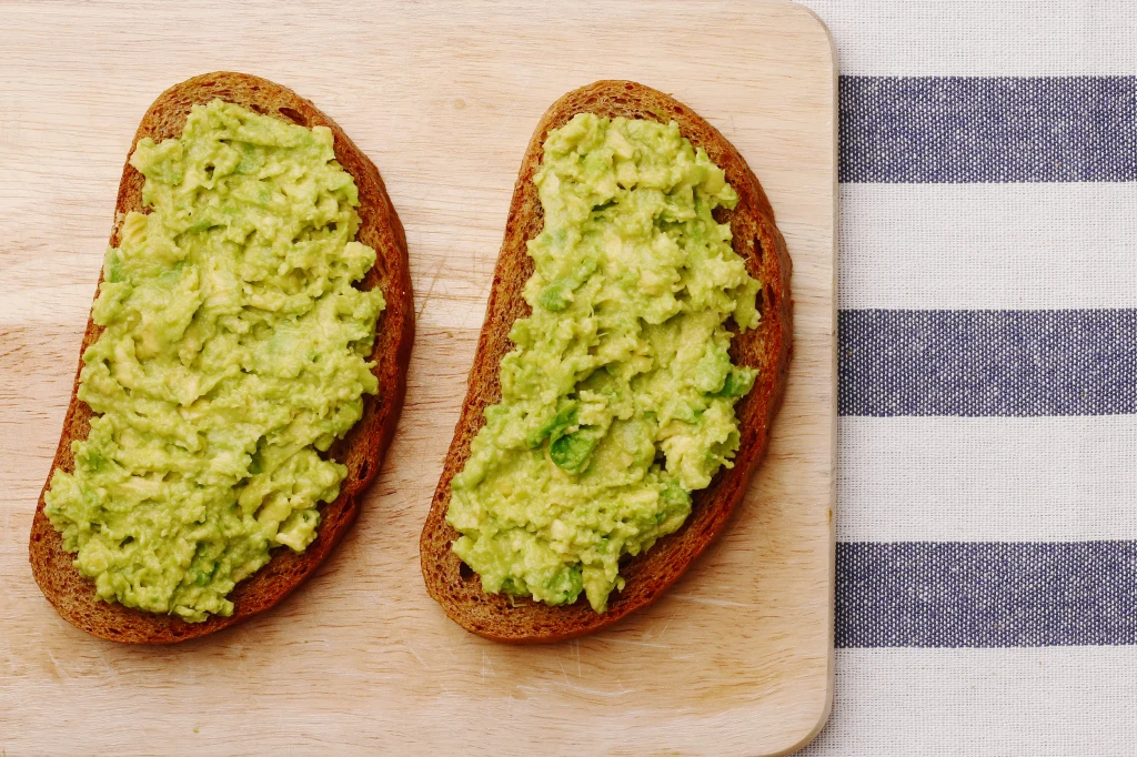 Dwie kromki chleba pełnoziarnistego posmarowane świeżo rozgniecionym awokado, ułożone na drewnianej desce kuchennej, obok białego obrusu w granatowe paski