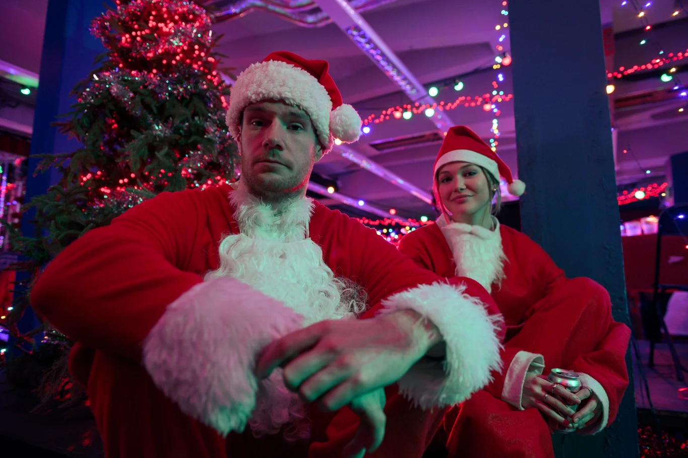 Mężczyzna i kobieta w strojach Mikołaja siedzący na tle udekorowanej świątecznie choinki oraz kolorowych lampek, w atmosferze nawiązującej do Bożego Narodzenia.