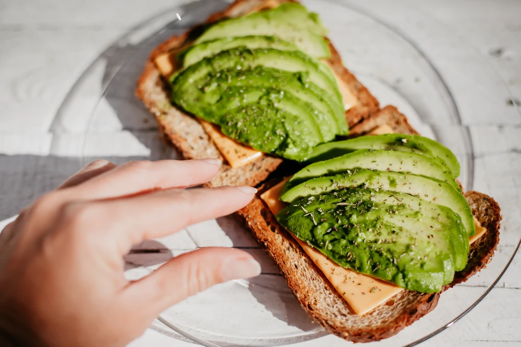 Dwie kromki chleba pełnoziarnistego z plastrem sera żółtego, plasterkami awokado oraz przyprawami, ułożone na szklanym talerzu, z ręką sięgającą po jedną z kanapek.
