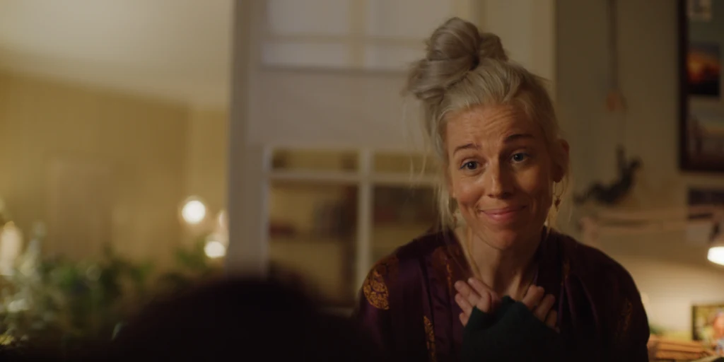 Kobieta o siwych włosach upiętych w kok stoi w ciepło oświetlonym wnętrzu mieszkania, trzymając ręce przy piersi i uśmiechając się życzliwie.