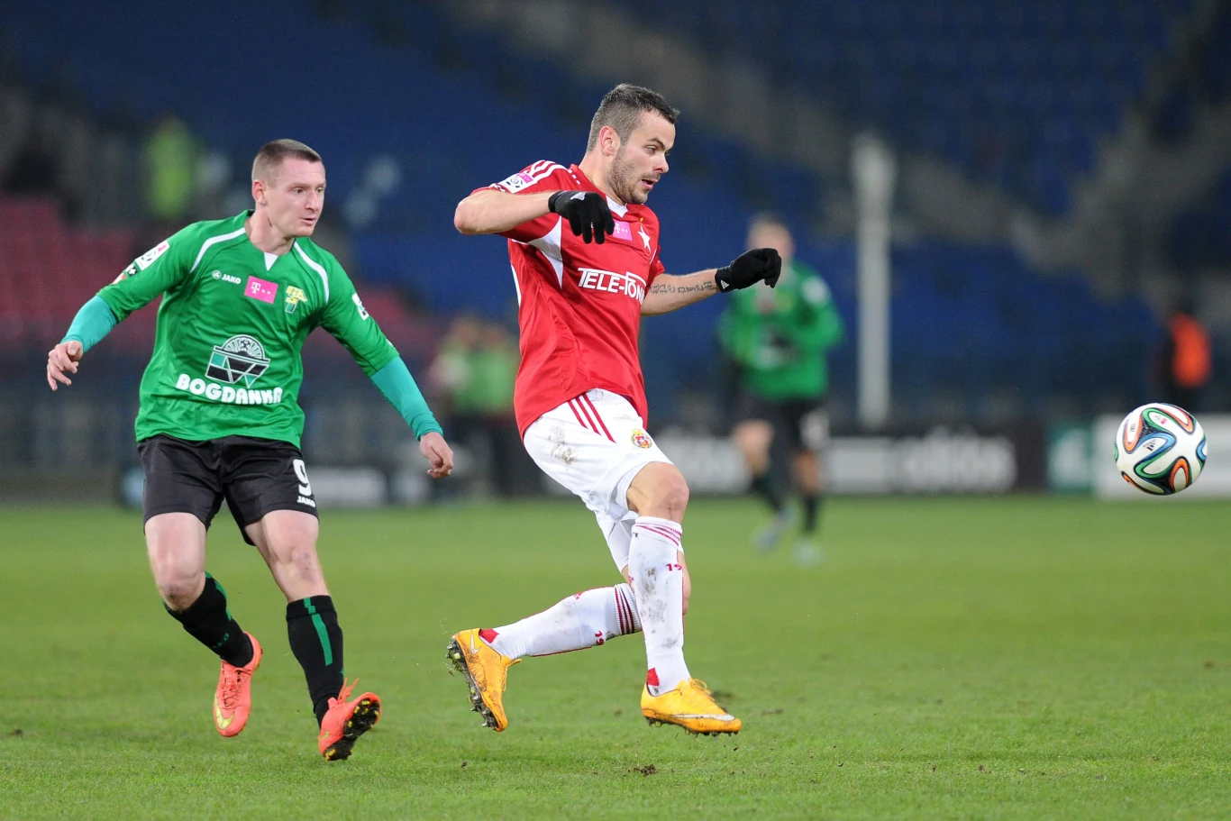 Dwóch piłkarzy w trakcie rywalizacji na boisku, jeden w zielonej koszulce próbuje odebrać piłkę zawodnikowi w czerwonym stroju, który jest przy piłce. W tle widoczni inni gracze i stadionowe trybuny.