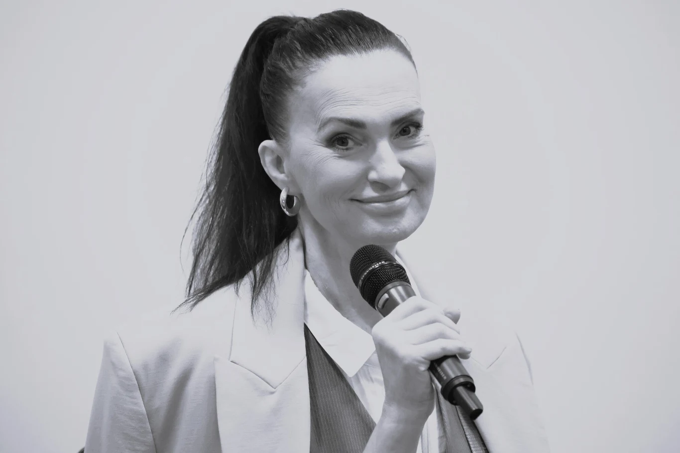 Kobieta o ciemnych włosach spiętych w kucyk uśmiecha się, trzymając mikrofon, ubrana w marynarkę i koszulę.