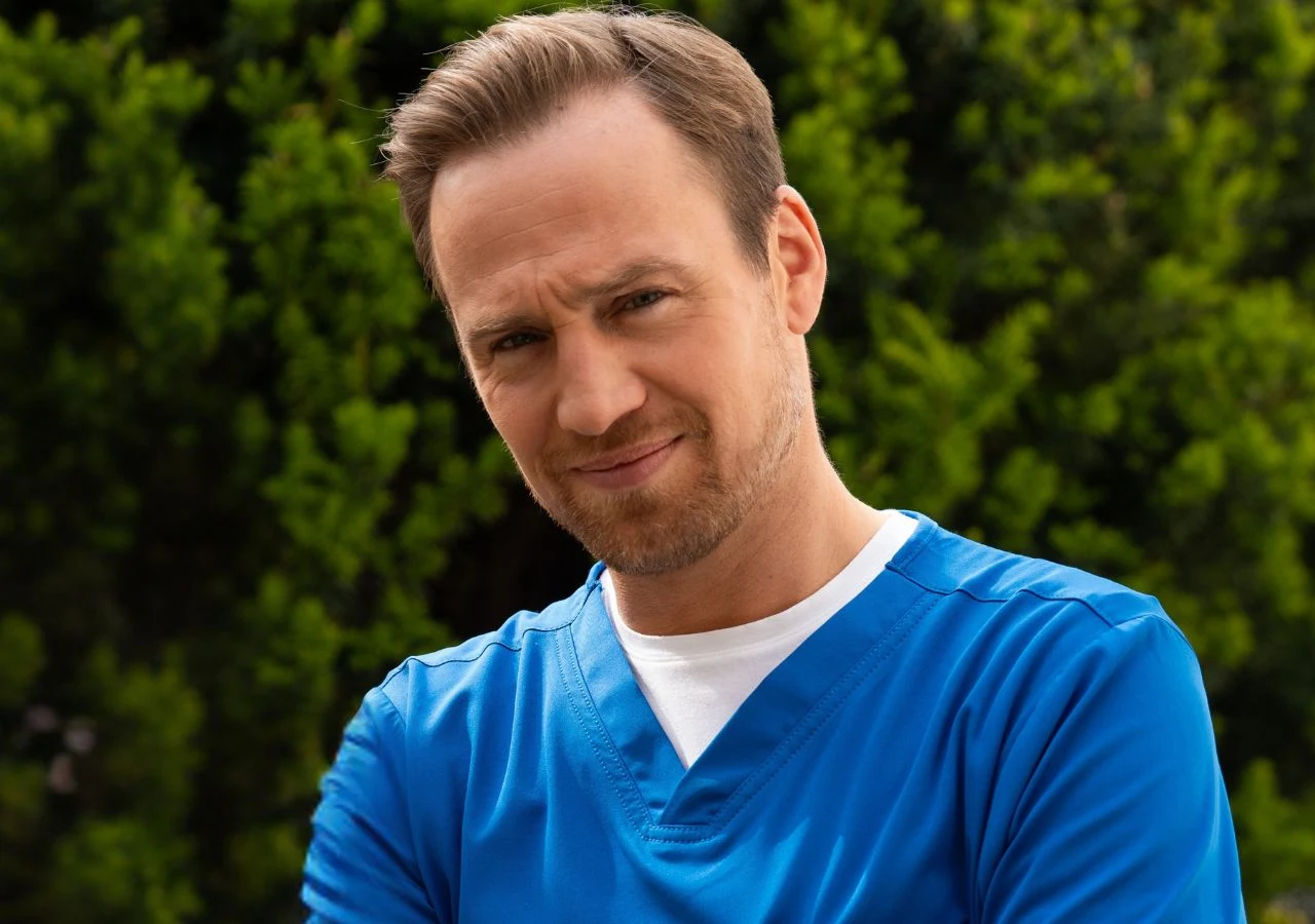 Mateusz Janicki na planie serialu "Na dobre i na złe" Mężczyzna w średnim wieku w niebieskim stroju medycznym stoi na tle zielonych krzewów, patrząc z lekkim uśmiechem i założonymi rękami.