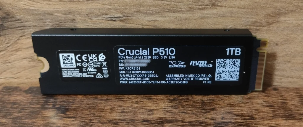 Crucial P510 Czarny dysk SSD NVMe Crucial P5 1TB z wyraźnie widocznymi oznaczeniami producenta i charakterystyką techniczną, umieszczony na drewnianej powierzchni.