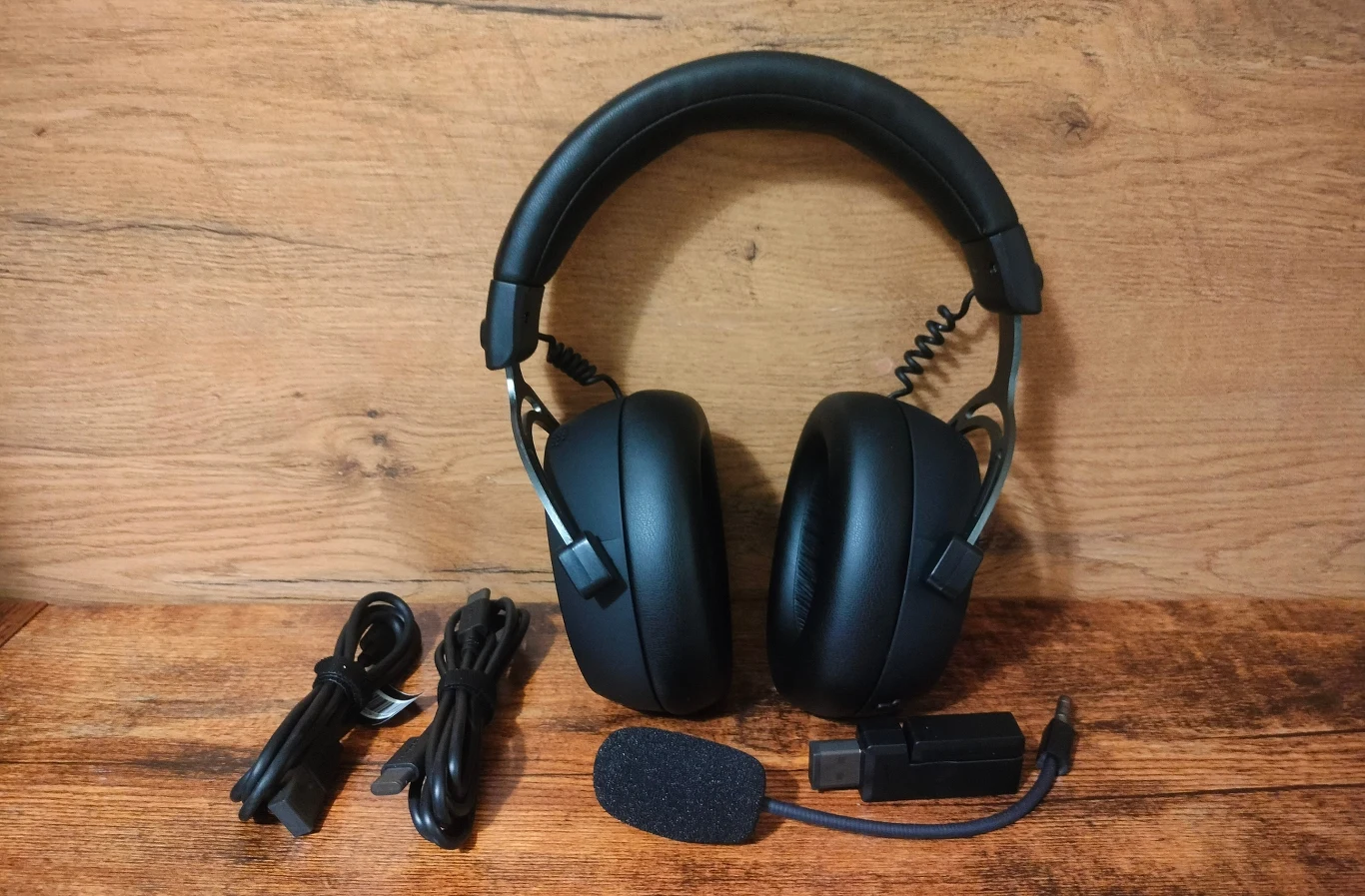 Hator Hypergang 3 Wireless Czarne słuchawki nauszne z miękkim pałąkiem, zestawem kabli, mikrofonem z gąbką i adapterem leżą na drewnianym blacie, w tle widoczna drewniana faktura ściany.