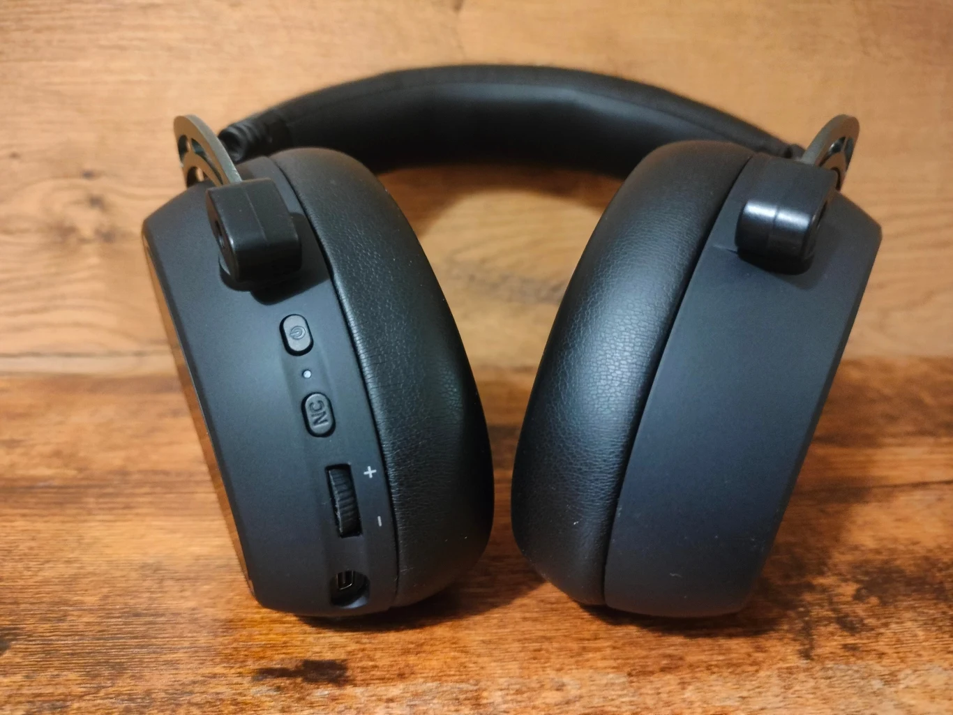 Hator Hypergang 3 Wireless Czarne bezprzewodowe słuchawki nauszne z miękkimi poduszkami i przyciskami sterującymi na jednej ze słuchawek, umieszczone na tle drewnianej powierzchni.