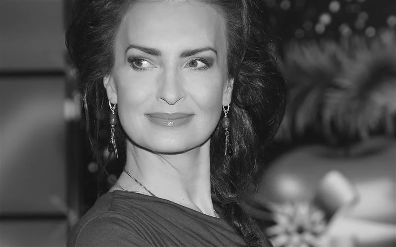 Kobieta z długimi, ciemnymi włosami spiętymi w warkocz, delikatny makijaż, długie kolczyki i łagodne światło wydobywające rysy twarzy, tło nieostre z elementami dekoracyjnymi.