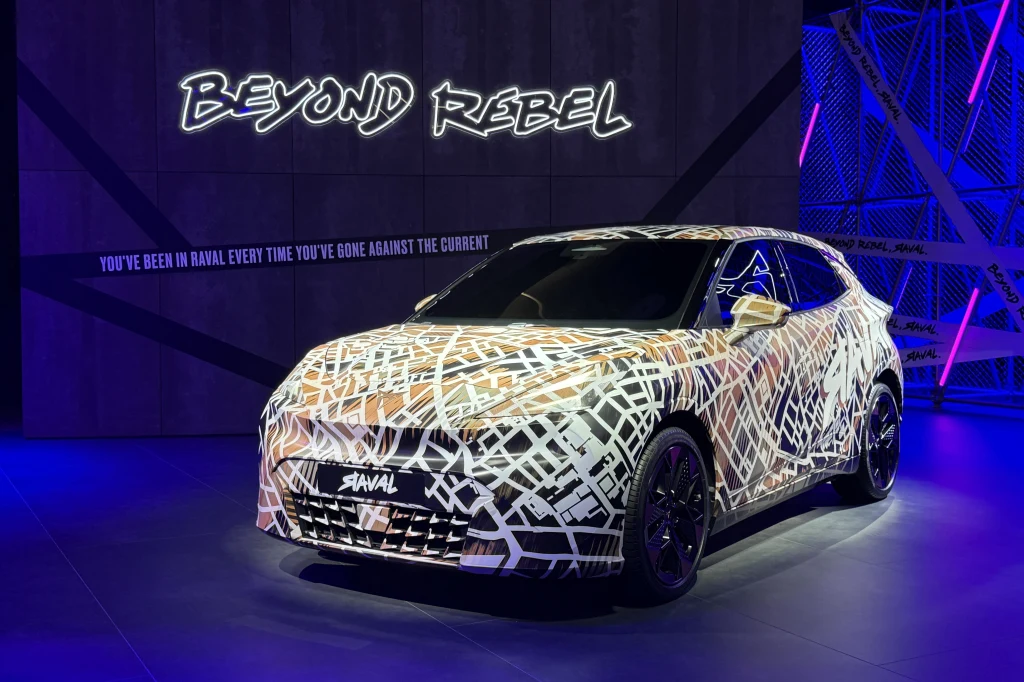 Cupra Raval - pierwsza z nowej generacji miejskich elektryków grupy Volkswagena. Nowoczesny samochód Haval pokryty unikalnym, graficznym wzorem, ustawiony pod jasnym oświetleniem na tle napisu 'BEYOND REBEL' oraz hasła promocyjnego, z dominującymi kolorami fioletu i niebieskiego.
