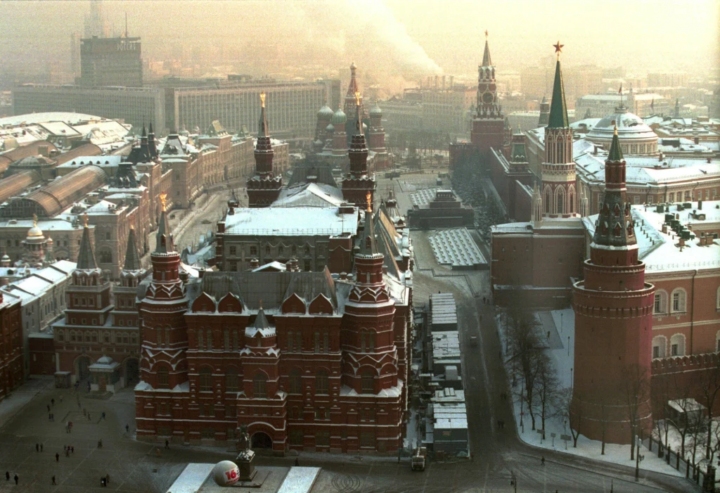 Zimowa panorama Placu Czerwonego w Moskwie z widocznymi murami Kremla oraz wieżami, zabytkowymi budynkami o czerwonych fasadach, ośnieżonymi dachami i miękkim światłem poranka unoszącym się nad miastem.
