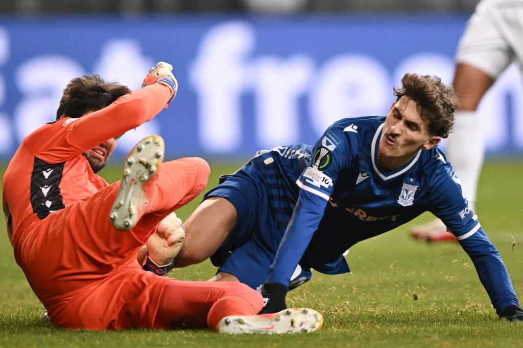 Intensywna sytuacja podczas meczu piłkarskiego, bramkarz w pomarańczowym stroju próbuje sięgnąć piłkę, podczas gdy zawodnik w niebieskim stroju z dużym wysiłkiem walczy o jej odzyskanie na murawie.