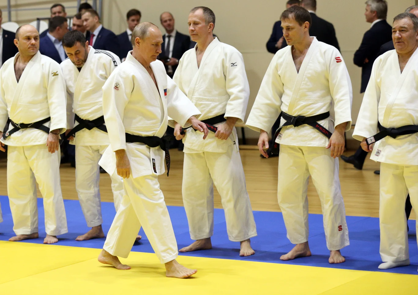 Putin w towarzystwie rosyjskich Judoków Grupa mężczyzn w białych judogach i z czarnymi pasami stoi na macie do judo. Jedna osoba idzie przed linią pozostałych, wszyscy skupieni na sytuacji przedstawionej na macie. W tle widoczne kilka osób w garniturach, prawdopodobnie obserwujących wydarzenie.