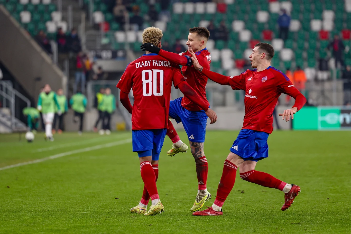 Zawodnicy drużyny piłkarskiej ubrani w czerwone koszulki i niebieskie spodenki świętują zdobycie bramki na boisku, jeden z nich ma numer 80 na plecach. W tle widać trybuny oraz innych piłkarzy.