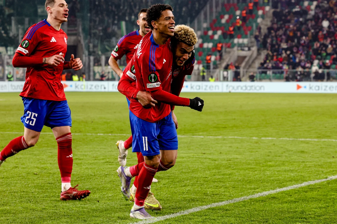 Grupa piłkarzy w czerwonych koszulkach świętuje zdobycie gola na boisku, w tle widać trybuny wypełnione kibicami.