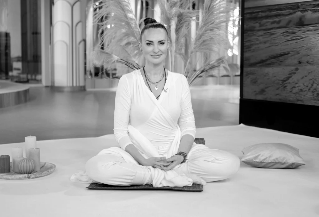 Agnieszka Maciąg uprawiała jogę Kobieta w białym stroju siedzi w pozycji medytacyjnej na macie, uśmiechnięta, w tle ozdobne rośliny i świece, obok poduszka i dekoracje, wnętrze utrzymane w spokojnych tonacjach.