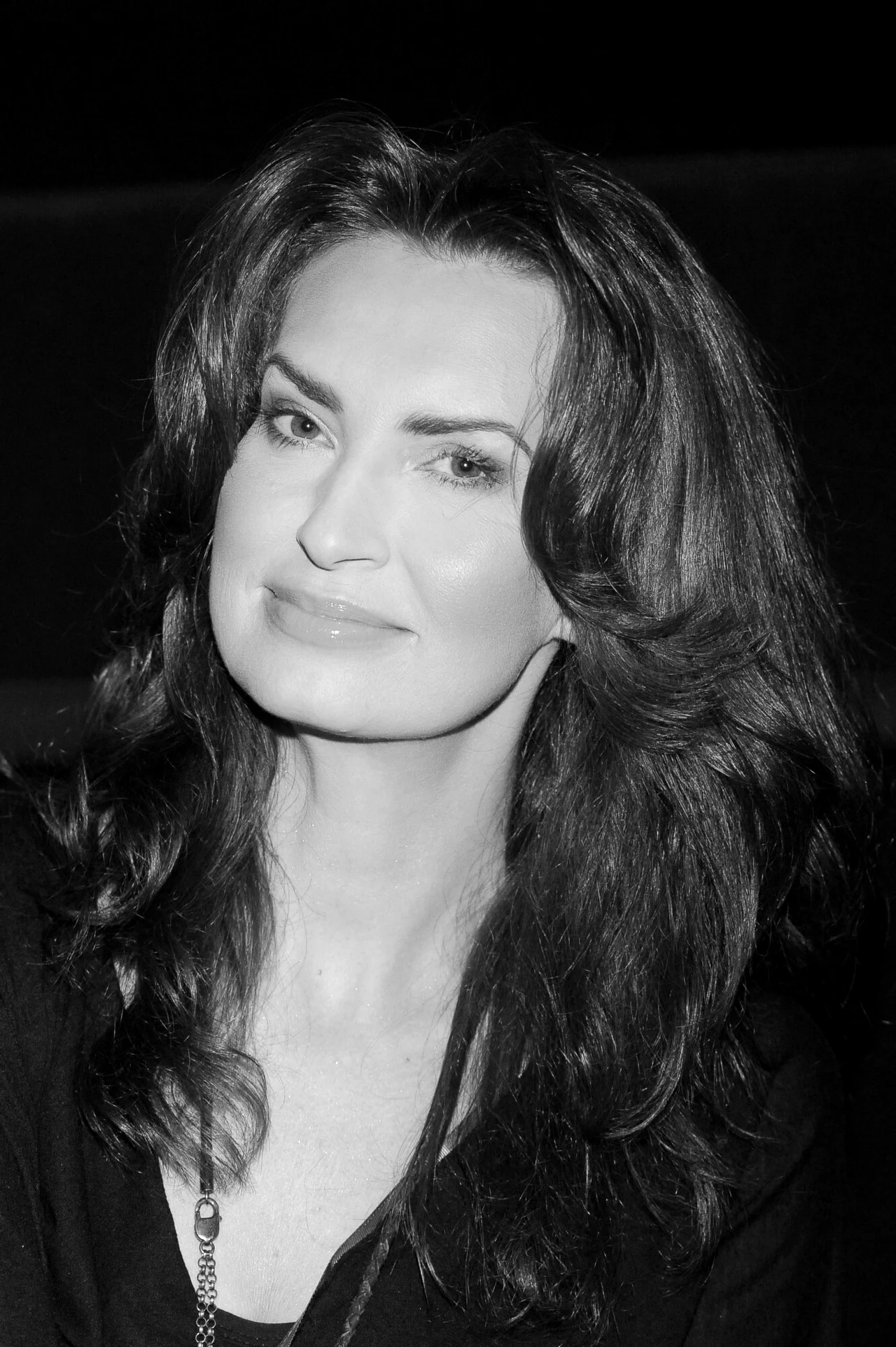 Kobieta o długich, ciemnych włosach z lekkim uśmiechem, patrząca w stronę obiektywu, sfotografowana w czerni i bieli.