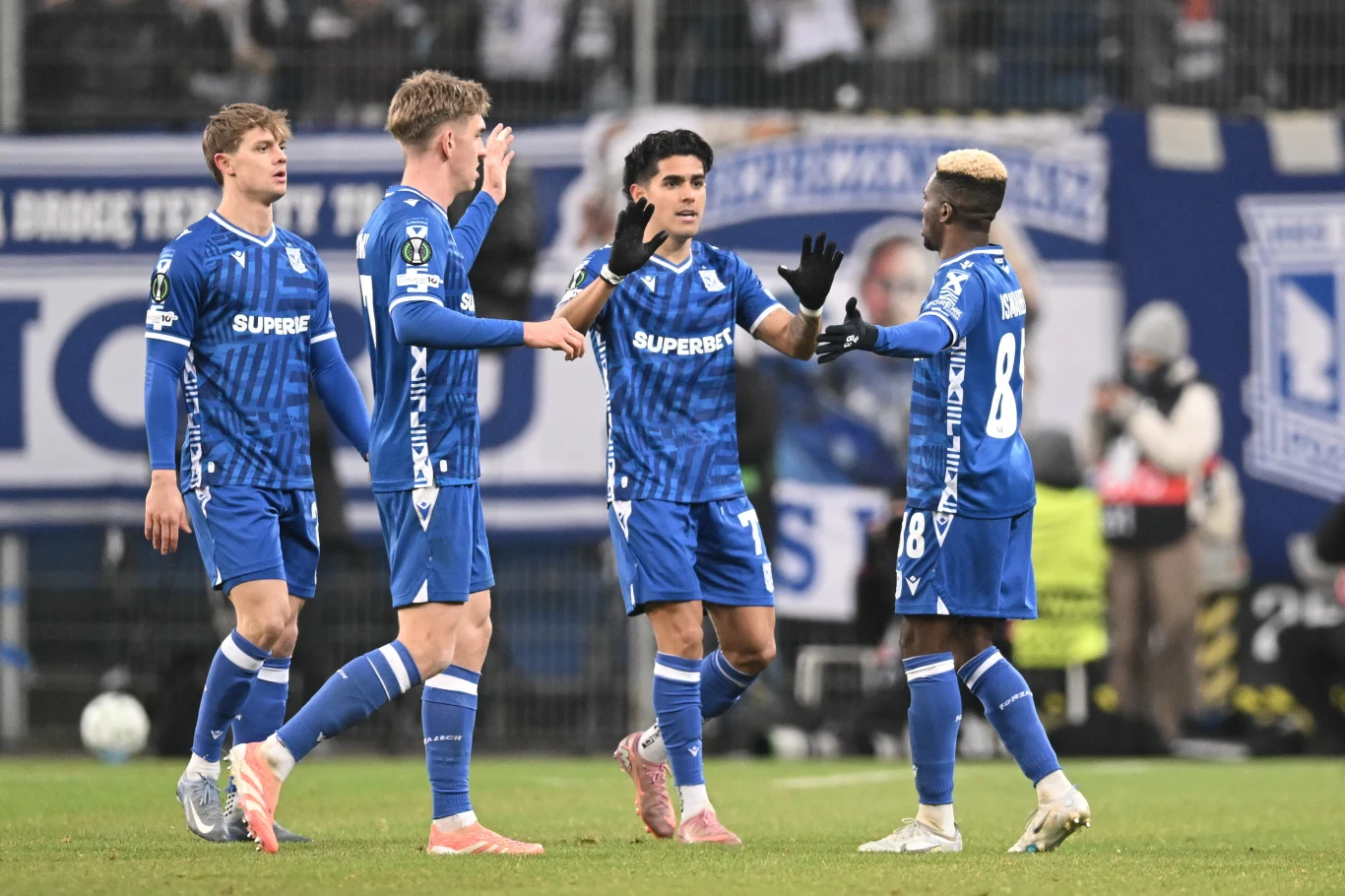 Czterech piłkarzy ubranych w niebieskie stroje drużyny podczas meczu na stadionie, wykonujących gesty gratulacji, w tle rozmazani kibice i elementy stadionowej infrastruktury.
