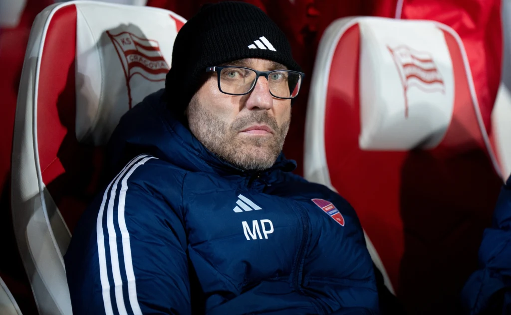 Mężczyzna w ciemnej kurtce sportowej oraz czapce z daszkiem i okularami siedzi na stadionowej ławce z czerwono-białymi siedzeniami, w tle widoczne są emblematy klubu.