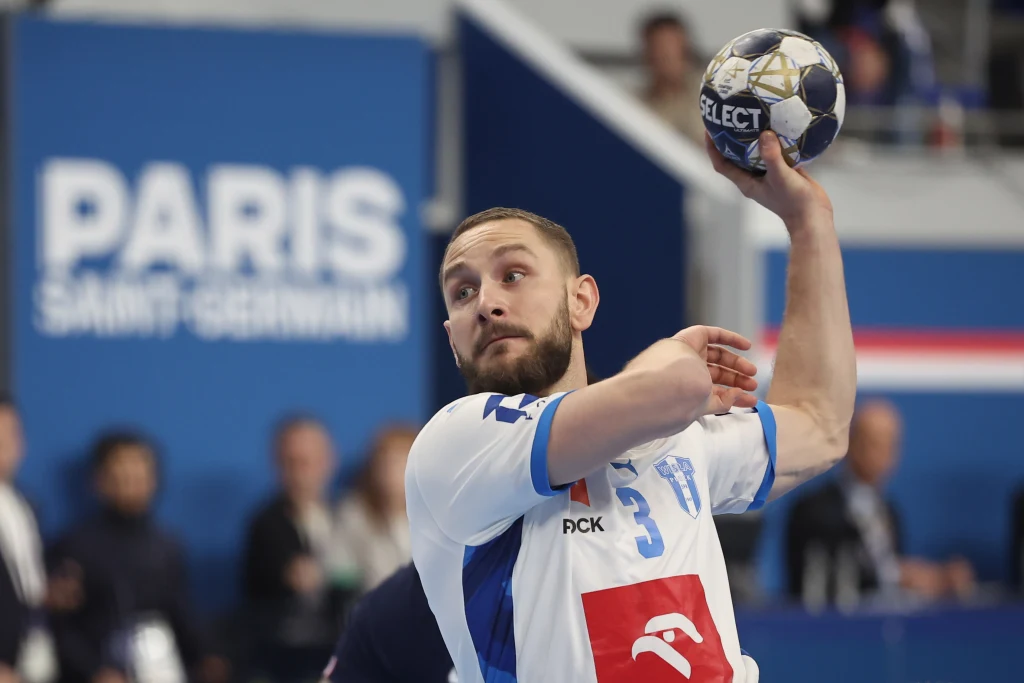 Zawodnik drużyny piłki ręcznej w trakcie gry, unoszący piłkę do rzutu, ujęty na tle niebieskiej hali z napisem Paris Saint Germain.