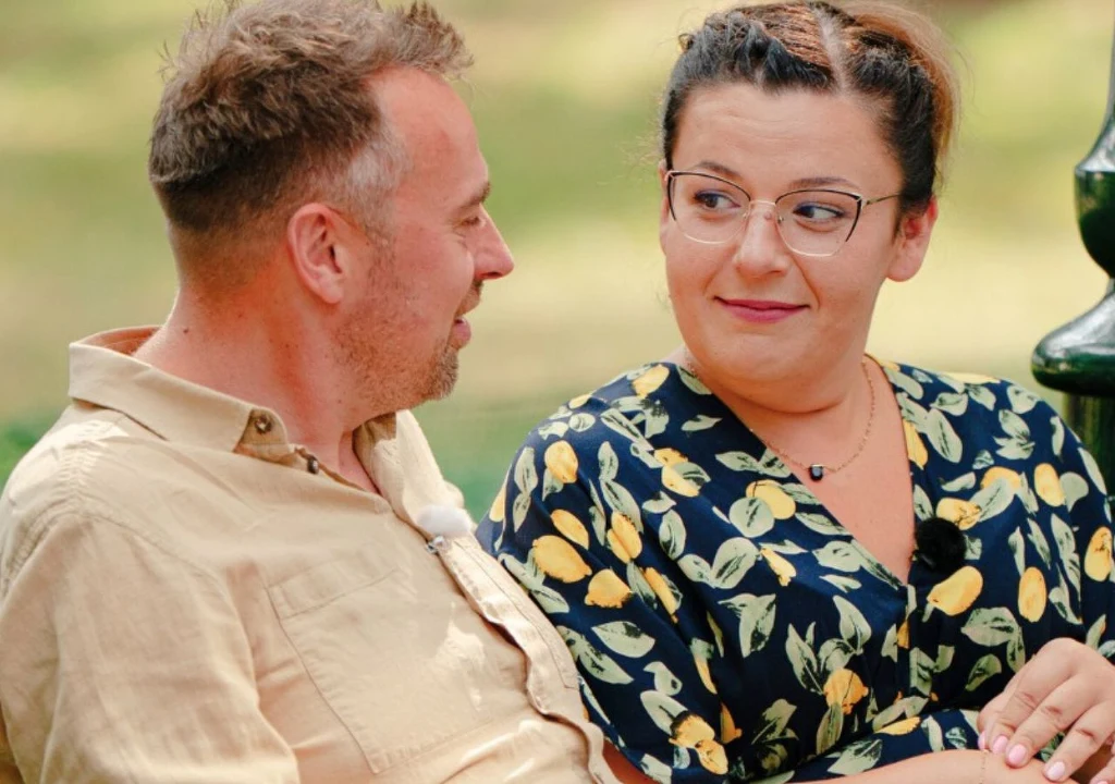 "Rolnik szuka żony": Barbara Boruch i Mateusz Uściłowski Dwoje ludzi siedzi na ławce w parku, uśmiechając się do siebie i prowadząc rozmowę. Kobieta nosi okulary i sukienkę w cytryny, mężczyzna ubrany jest w jasną koszulę.