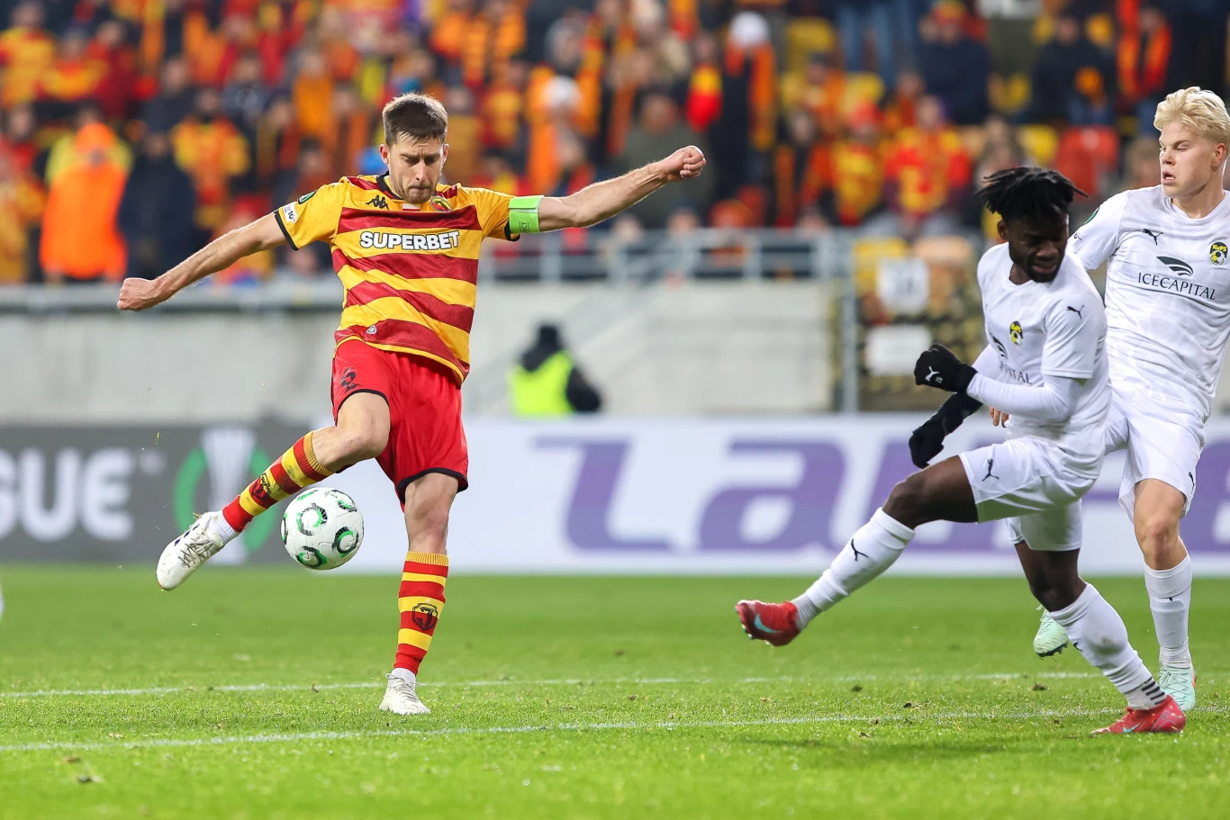 Piłkarz w żółto-czerwonej koszulce wykonuje kopnięcie piłki podczas meczu, wokół niego dwaj zawodnicy w białych strojach próbują go zablokować, w tle widoczna trybuna z kibicami.