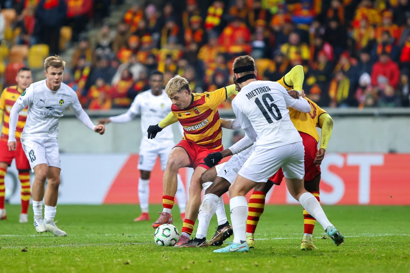 Zawodnicy dwóch drużyn piłkarskich rywalizują o piłkę w trakcie meczu na stadionie, w tle widać trybuny z kibicami ubranymi w barwy klubowe.