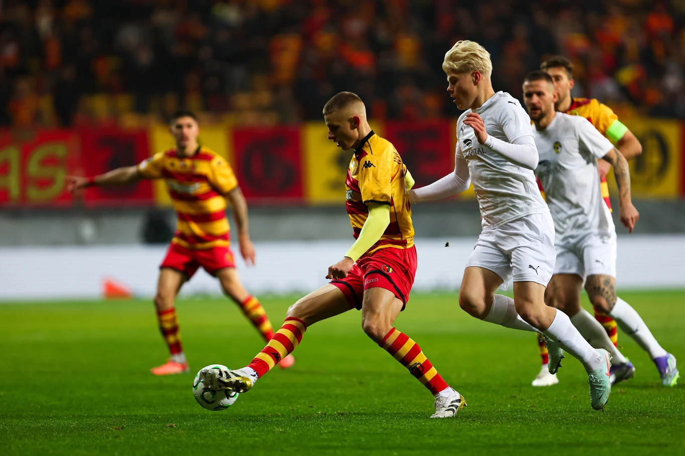 Piłkarze dwóch drużyn walczą o piłkę na boisku w świetle stadionowych jupiterów, w tle widać kibiców oraz kolorowe flagi i transparenty, zawodnicy noszą stroje w barwach żółto-czerwonych oraz białych.