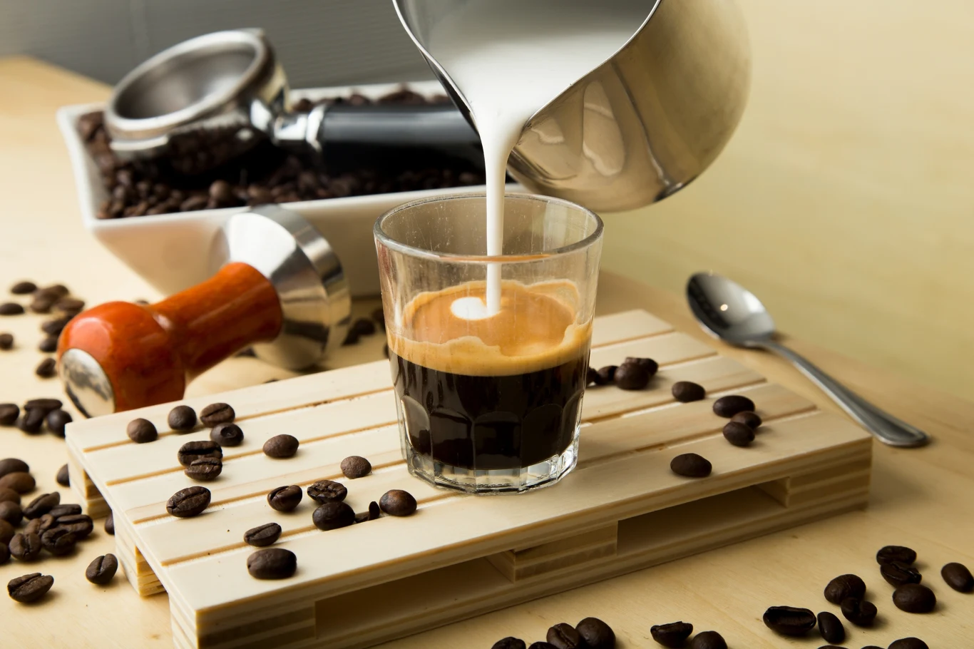 Świeżo zaparzona kawa espresso w szklance, do której wlewane jest mleko. W tle ziarna kawy, tamper i pojemnik z ziarnami umieszczony na drewnianym stole.