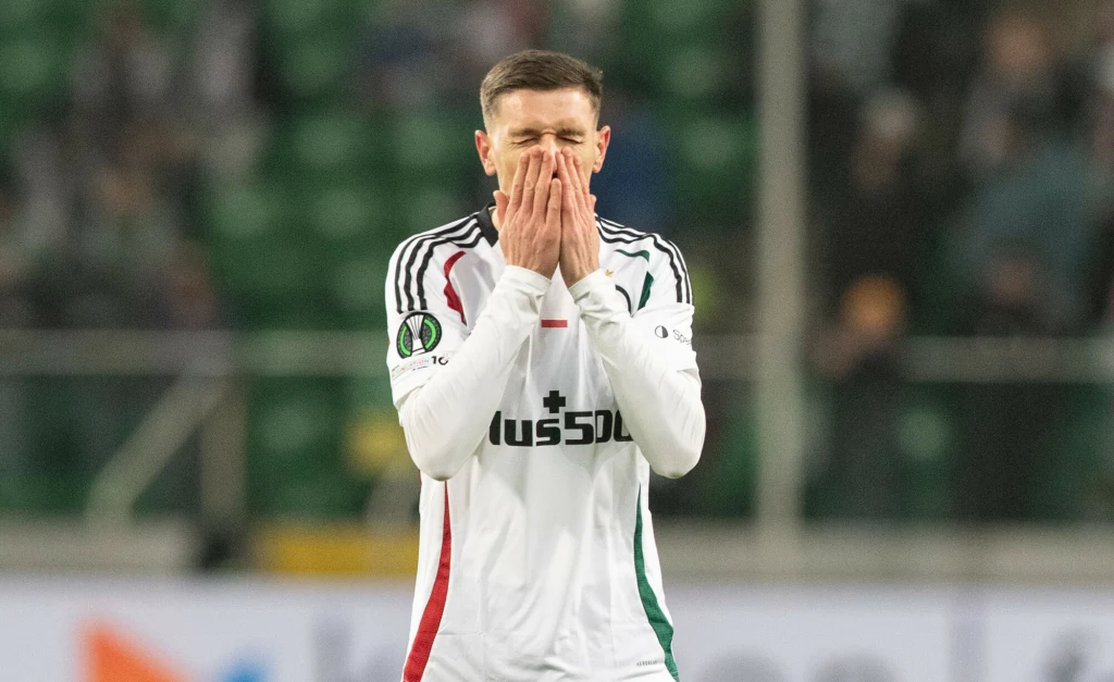 Piłkarz w białym stroju z logo sponsora i naszywkami klubowymi zasłania twarz dłońmi w geście rozczarowania. W tle rozmazana widownia stadionu.