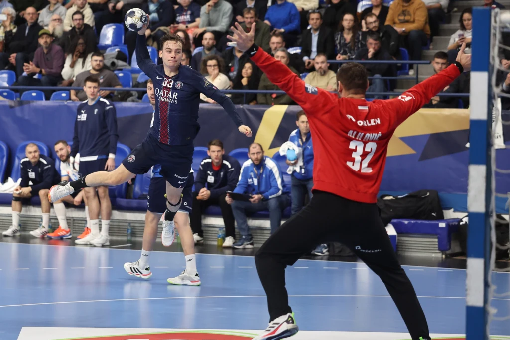 Zawodnik drużyny PSG wykonuje rzut podczas meczu piłki ręcznej, próbując zdobyć bramkę, podczas gdy bramkarz drużyny przeciwnej stara się obronić. W tle widoczna ławka rezerwowych oraz widownia skupiona na przebiegu akcji.