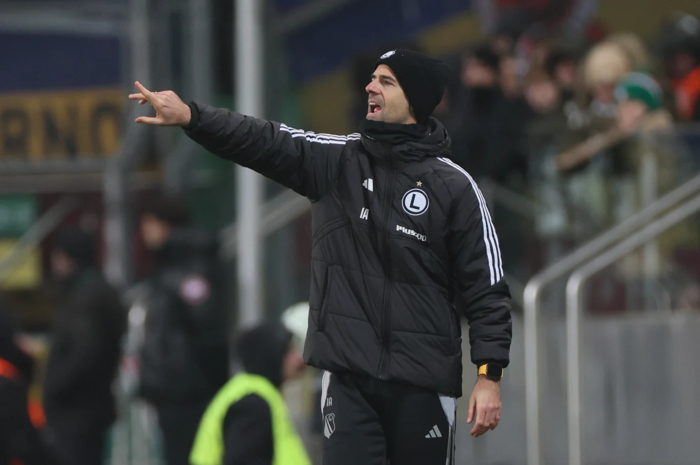 Trener drużyny piłkarskiej Legii Warszawa w sportowej odzieży, energicznie gestykuluje na linii bocznej boiska podczas meczu, w tle rozmazani kibice i osoby obsługi technicznej.