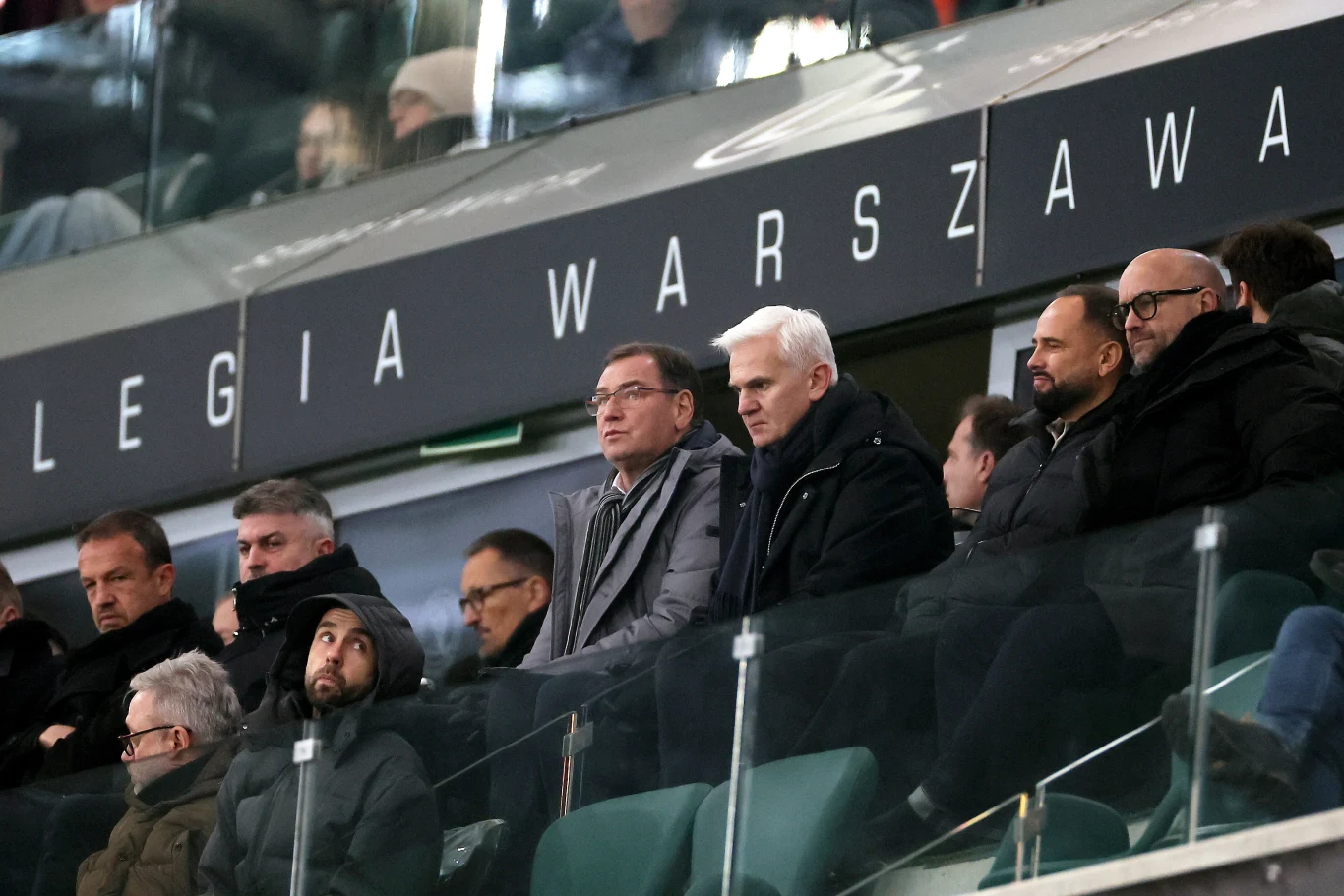Grupa mężczyzn w eleganckich ubraniach siedzi na trybunach stadionu piłkarskiego Legii Warszawa, za taflą szkła, skupiona na wydarzeniach na boisku. W tle widoczny napis 'Legia Warszawa'.