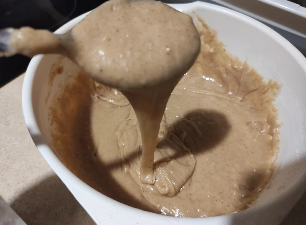 Gęste, kremowe ciasto o jasnobrązowej barwie przelewane z łyżki do dużej białej miski, konsystencja wskazuje na masę do wypieków.