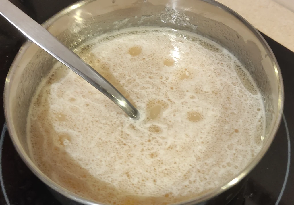 Garnek wypełniony gotującą się owsianką, z wyraźnie widoczną pianą na powierzchni. W garnku znajduje się metalowa łyżka mieszająca zawartość. Naczynie stoi na płycie grzewczej.