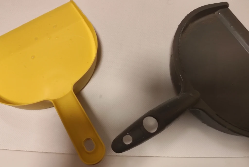 Aby trik zadziałał potrzebujesz zmiotki z uchwytem w kształcie rynny Dwie szufelki do zmiatania, jedna w kolorze żółtym, druga czarna, umieszczone na jasnym tle, uchwyty skierowane w tę samą stronę.