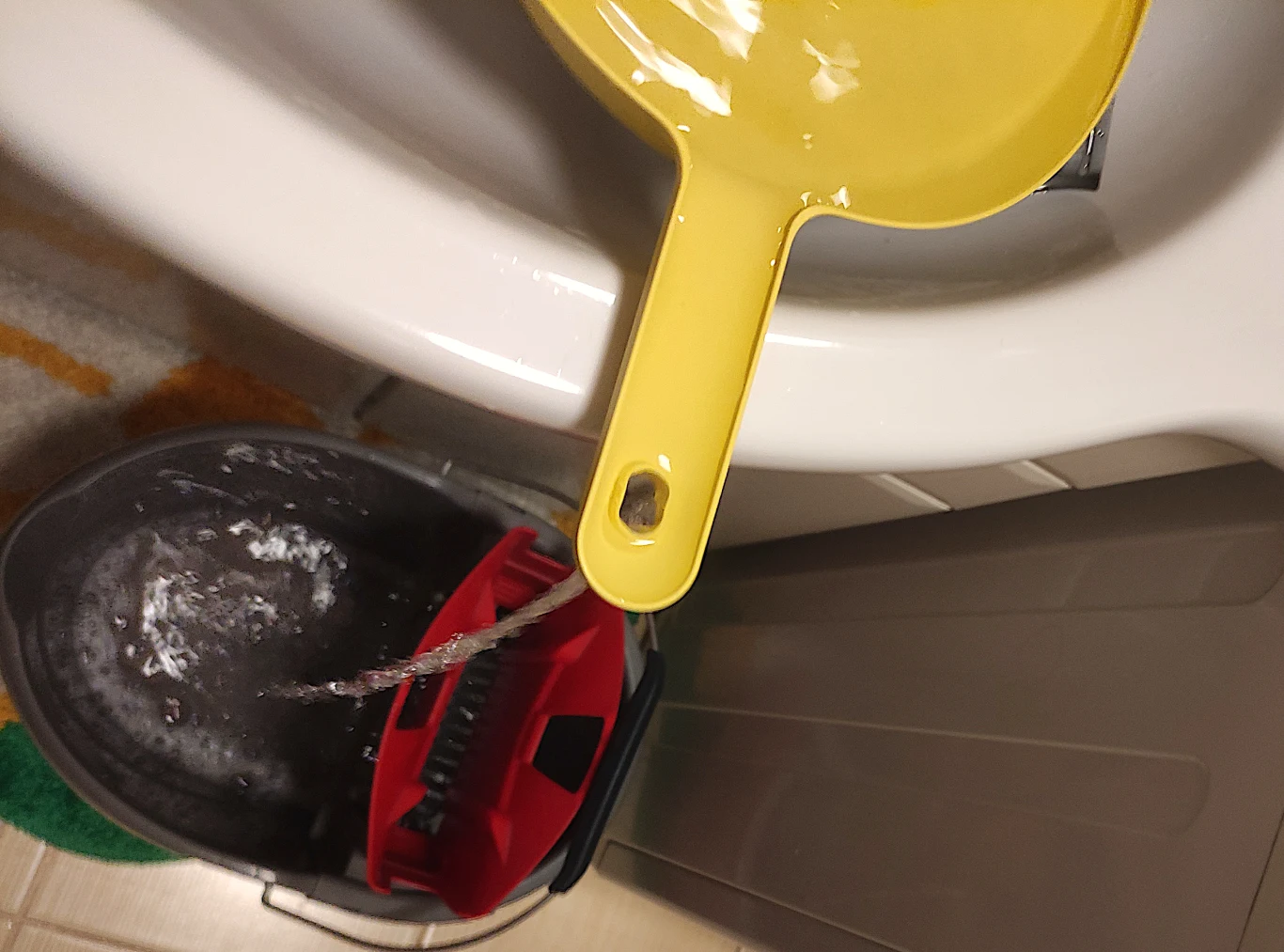 Otwór w uchwycie zmiotki nie był przeszkodą w napełnieniu wiadra wodą Żółta szufelka oparta na białej misce lub umywalce, wylewająca wodę do plastikowego wiadra z mopem i pianą. Scena kojarzy się z czynnością sprzątania lub wylewania brudnej wody po myciu.