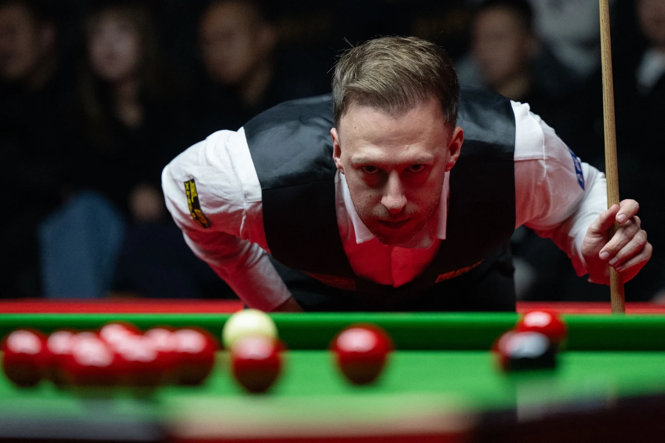 Zawodnik snookera w skupieniu przygotowuje się do wykonania uderzenia, celując kijem bilardowym w białą bilę otoczoną kolorowymi bilami, rozgrywka toczy się przy stole bilardowym, w tle obecni widzowie.